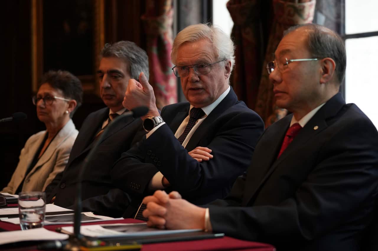 Four people in formal attire sit at a long table during a meeting, with a white-haired man in glasses near the center gesturing with a raised fist while speaking.