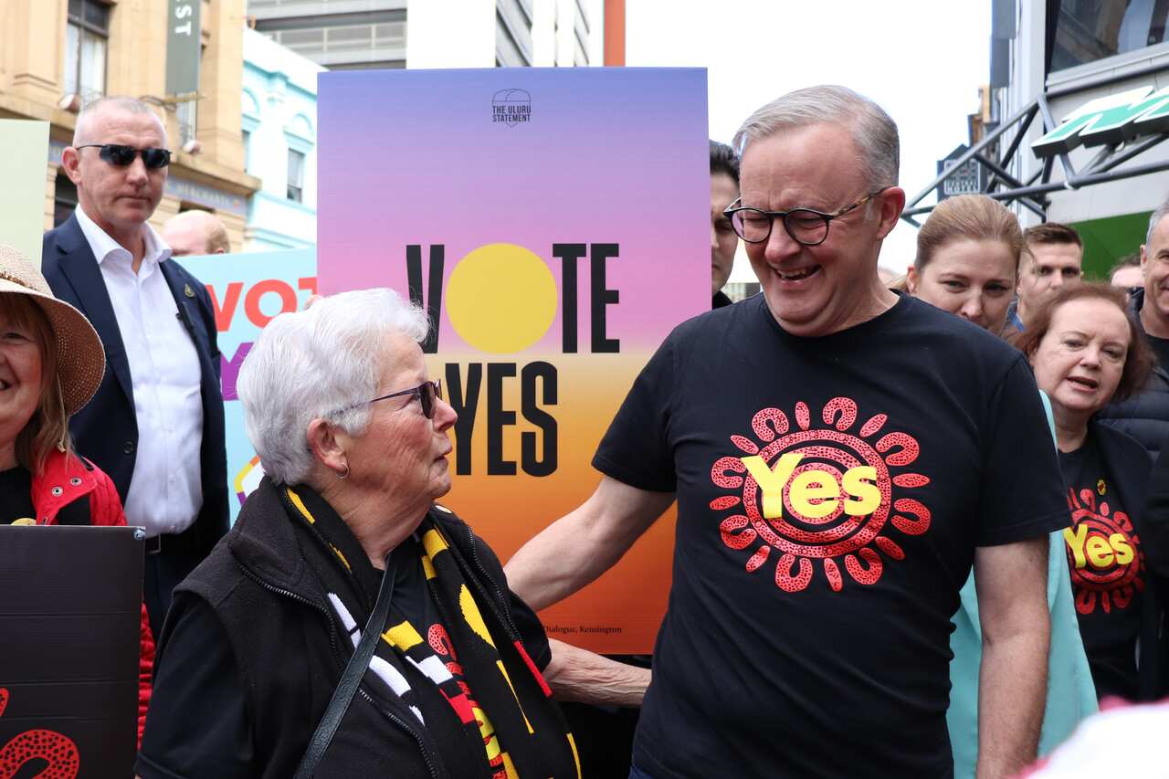 Anthony Albanese wearing a black t-shirt with the word Yes written in yellow on the front. He is standing next to a woman outside. There is a purple sign behind them with the words Vote Yes written on it.