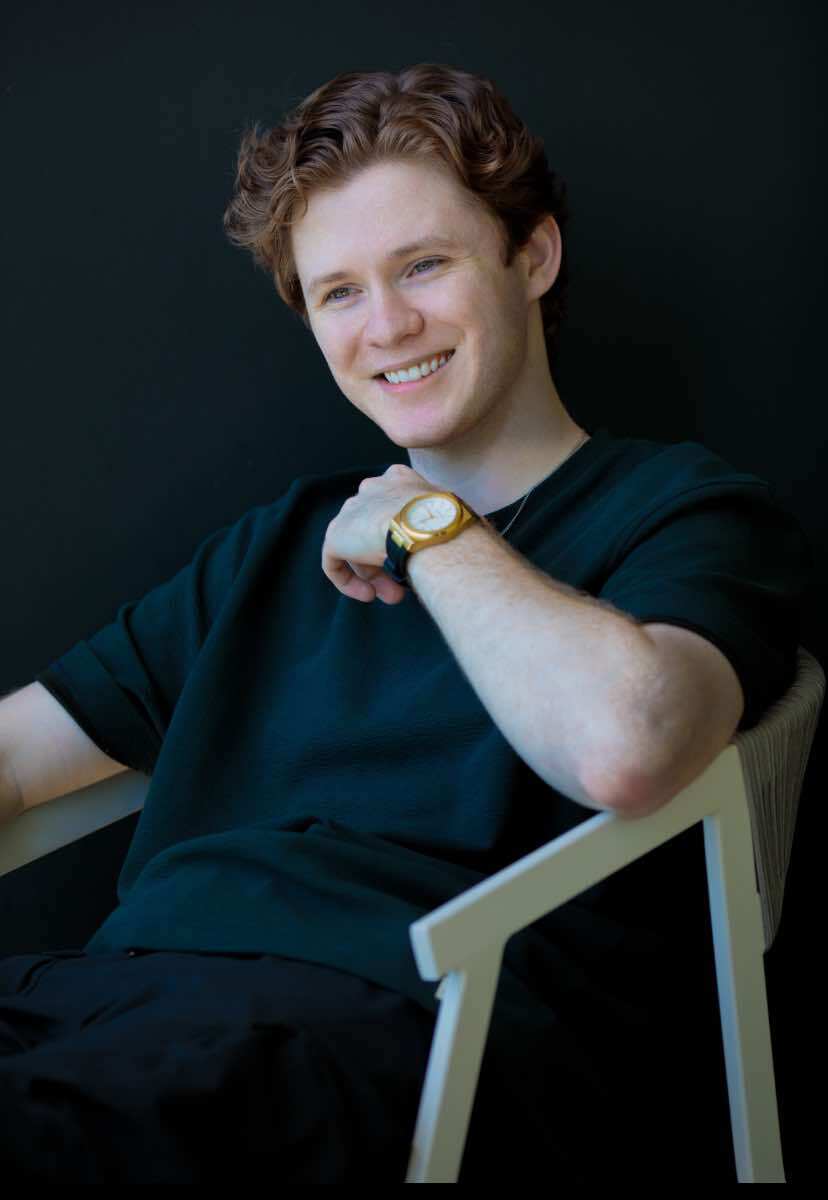 A young man wearing a dark green T-shirt sits back in a white, plastic chair. 