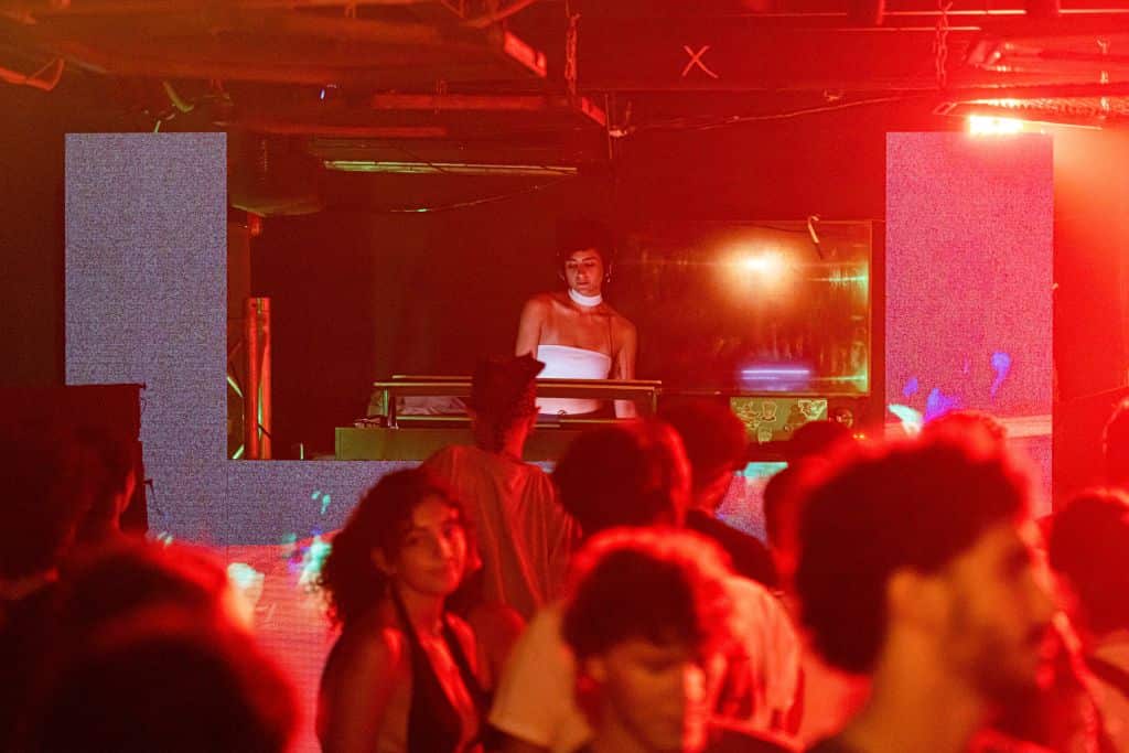 A woman plays a DJ set at a venue as people dance in front of her. 