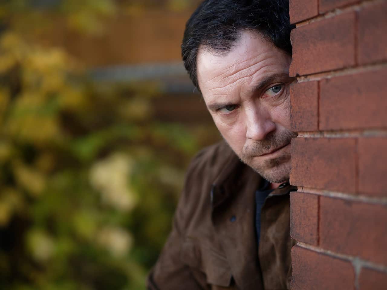 A man in a brown leather jacket peers around the edge of a brick wall.