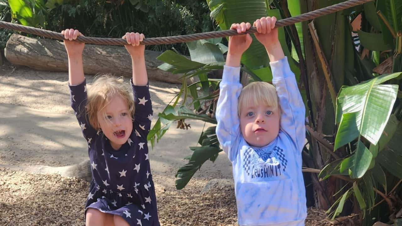 two young blonde children a boy and girl swinging on a rope in a park