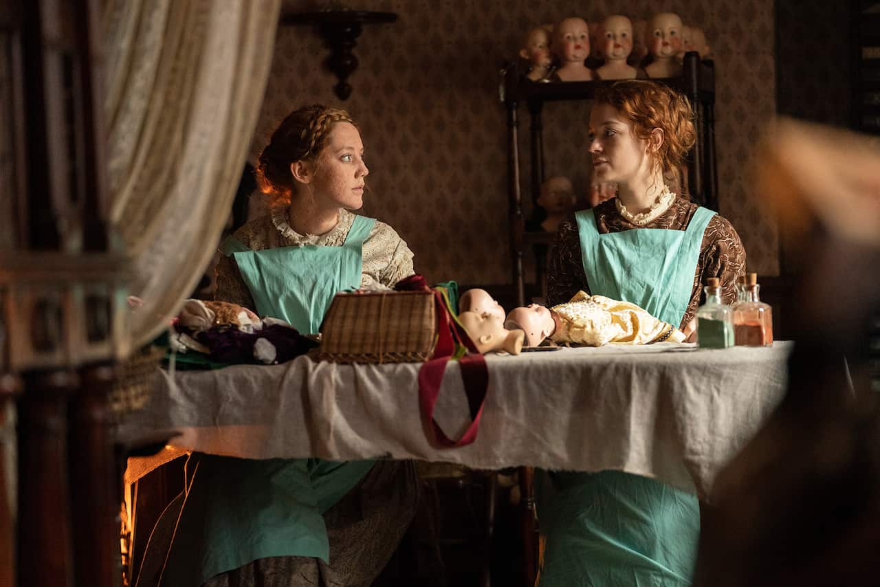 Two young women with red hair, both wearing aprons over their dresses, sit at a table. 