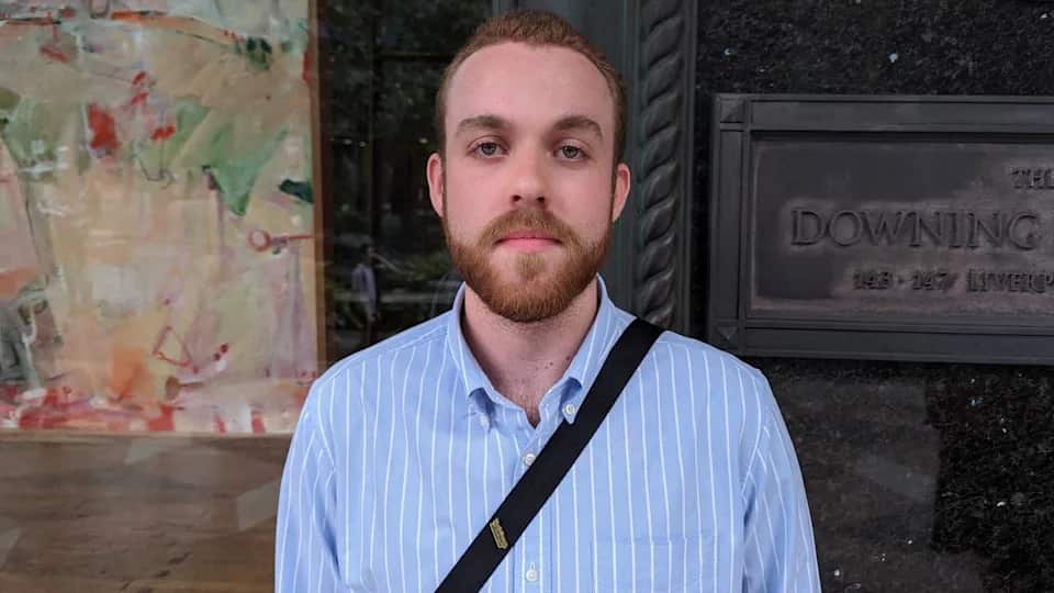 A young white man with short red hair and a beard, wearing a striped blue shirt