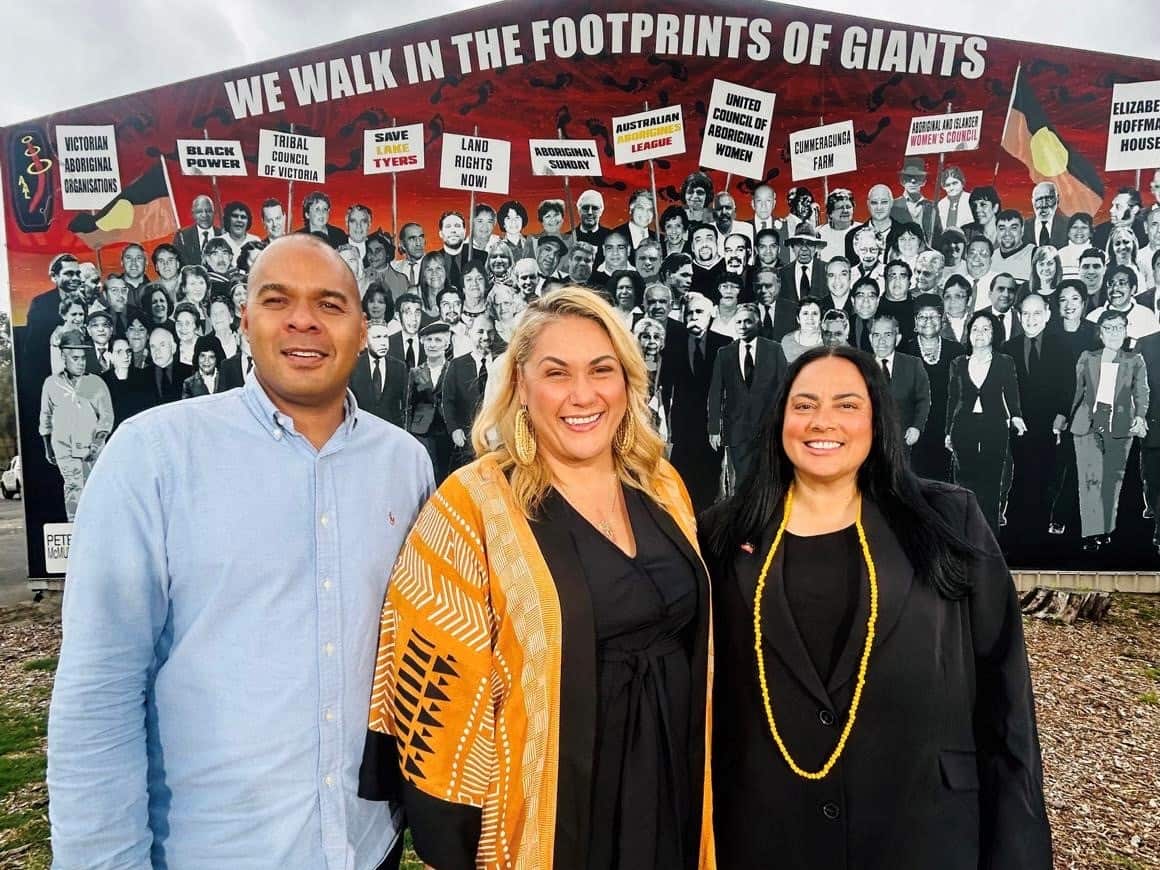 A man and two women standing near each other and smiling, in front of a wall that has a mural of people marching with signs displaying First Nations messages.