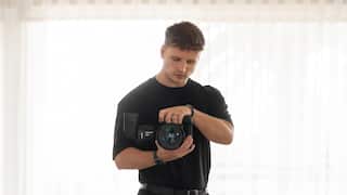 a young blonde man holds a camera while standing in front of white curtains