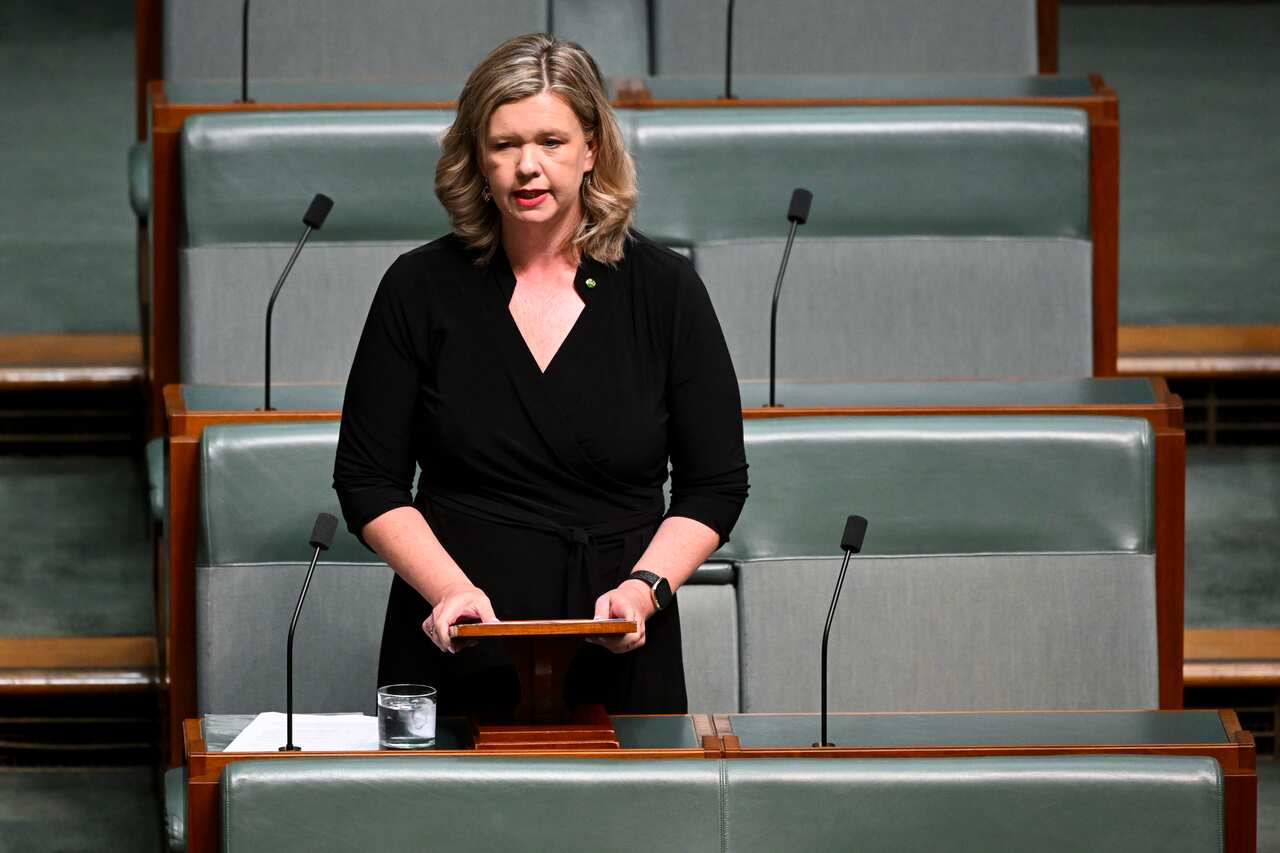 A woman wearing a black dress standing and speaking in front of microphones.