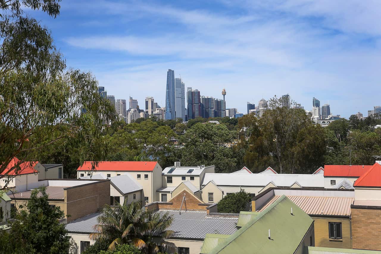 A view over suburban rooftops and lush green trees reveals the Sydney skyline in the distance, featuring prominent landmarks like Crown Sydney and the Sydney Tower Eye under a bright, partly cloudy sky.