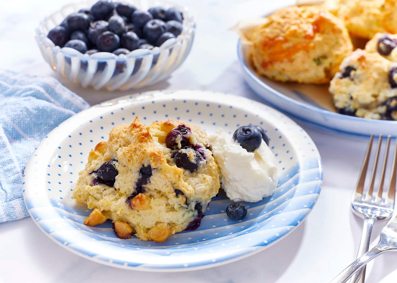 A shallow blue and white bowl with a spot and stripe pattern holds a rough-shaped baked scone-like biscuit. A scoop of cream and a few blueberries sit alongside, in the bowl. A tray can be seen in the background with more biscuits. 