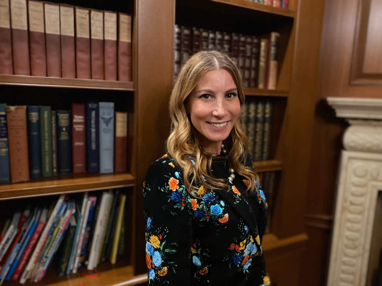 A woman in a dark dress with colourful flowers on it standing in front of a book case