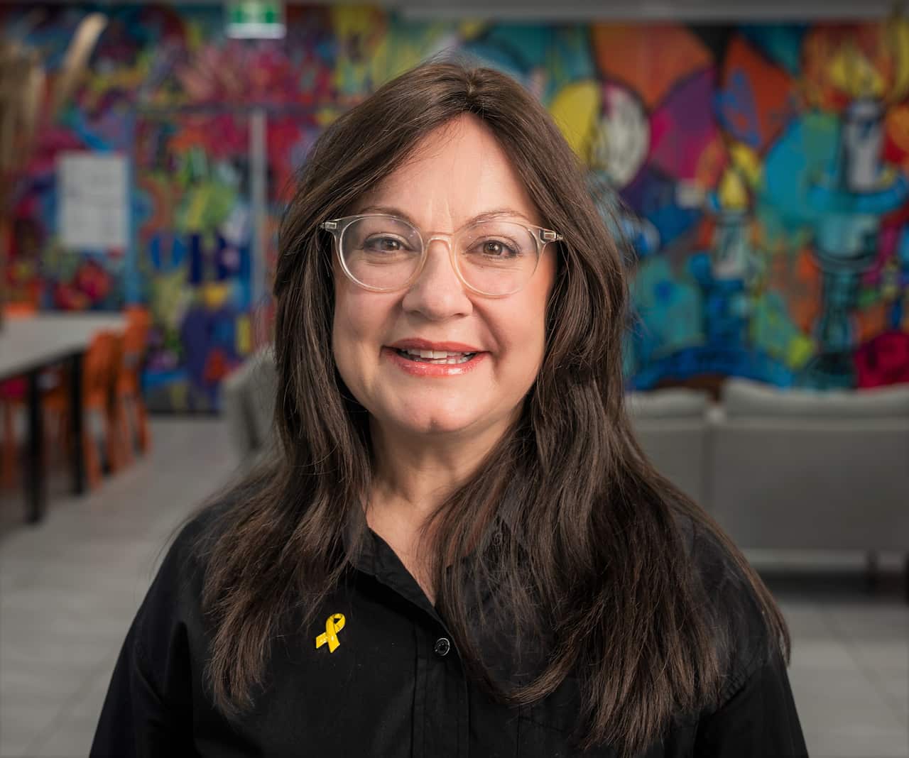 A woman with long brown hair and wearing glasses sits in front of a colourful mural wall.