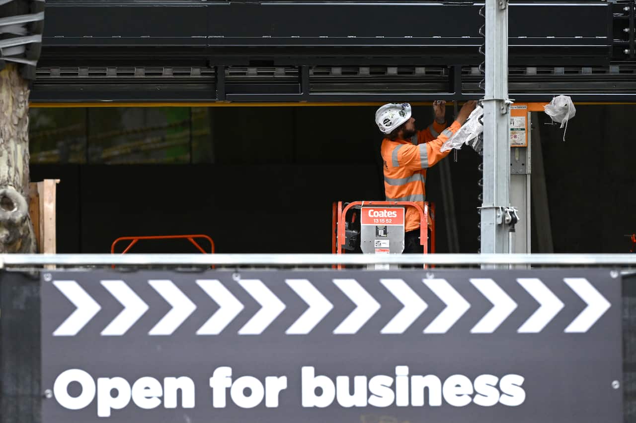 Man in construction uniform on machinery wearing a helmet. Behind him is a construction site. In front of him is a sign that says 'open for business'.