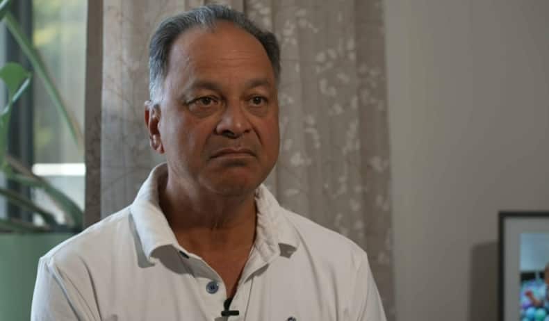 An Indian Australian man wearing a white collared shirt sits in a living room.