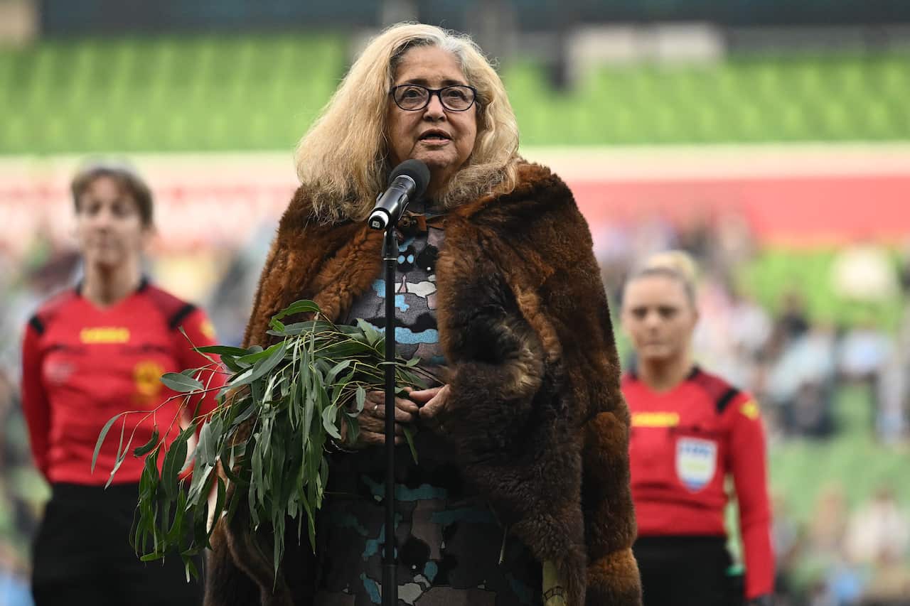 A woman wearing a fur cloak and holding a branch of gum leaves stands behind a microphone outside