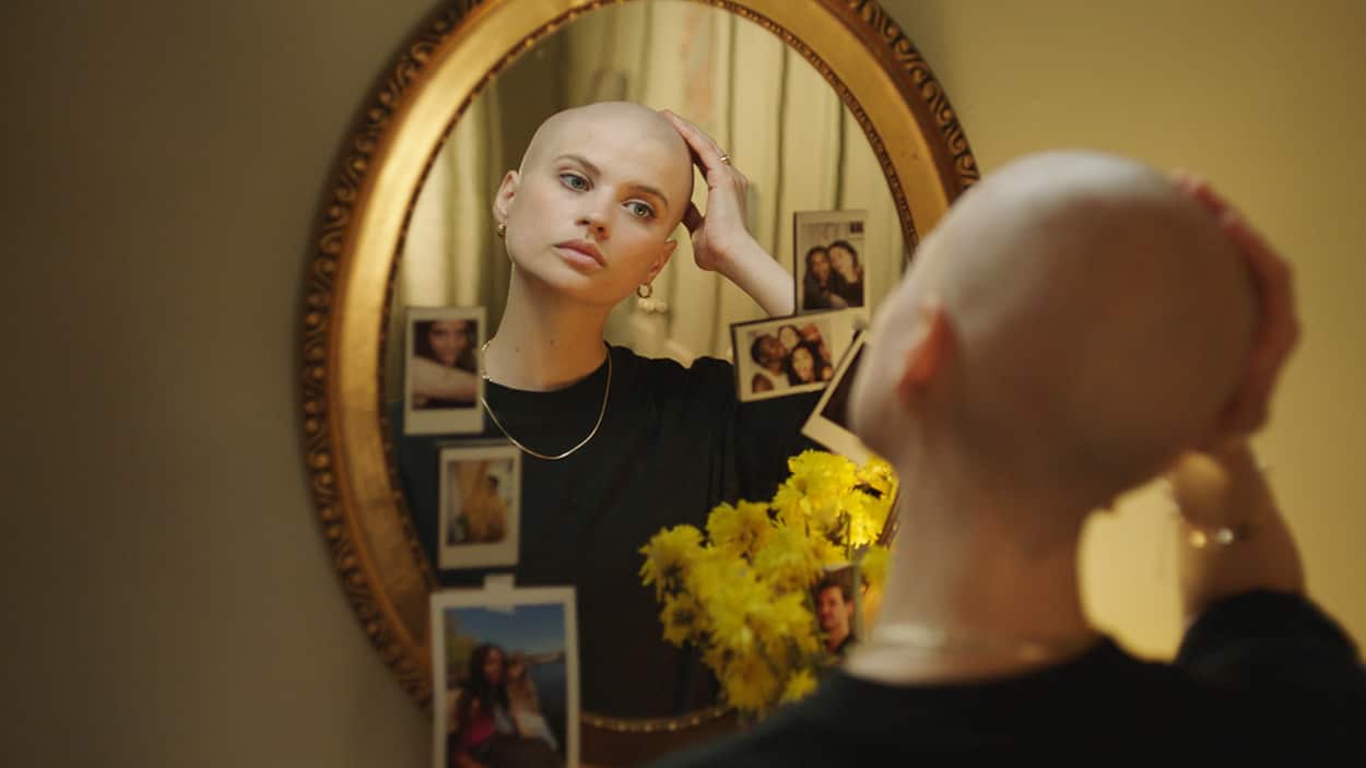 A young woman looks at herself in the mirror and touches her shaved head.jpg