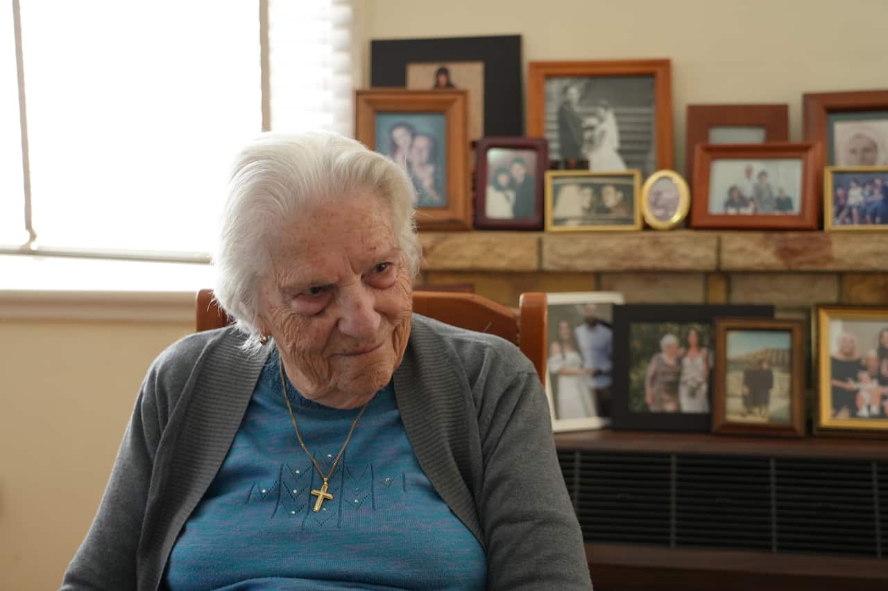An older woman with white hair, wearing a blue shirt and grey cardigan, with a gold cross around her neck, in the background, out-of-focus family photos.