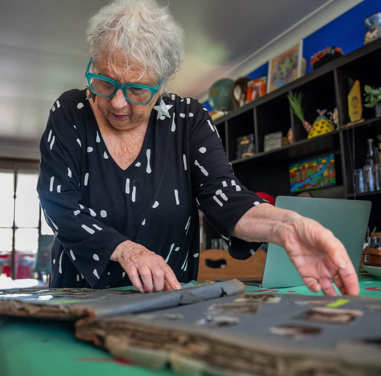 A woman in a black dress stands over a table looking at old photos. 
