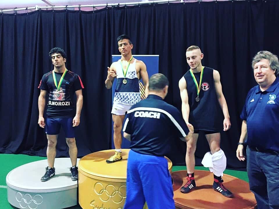 Three men on a podium, the one winning gold gesturing to the camera