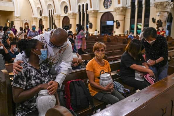 A priest comforts a woman sitting in a church pew and holding an urn