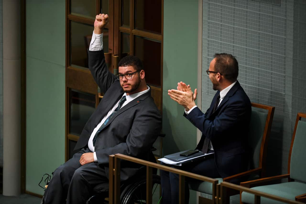 Jordon raises his fist in the air, sitting in his wheelchair while Adam Bandt applauds him.