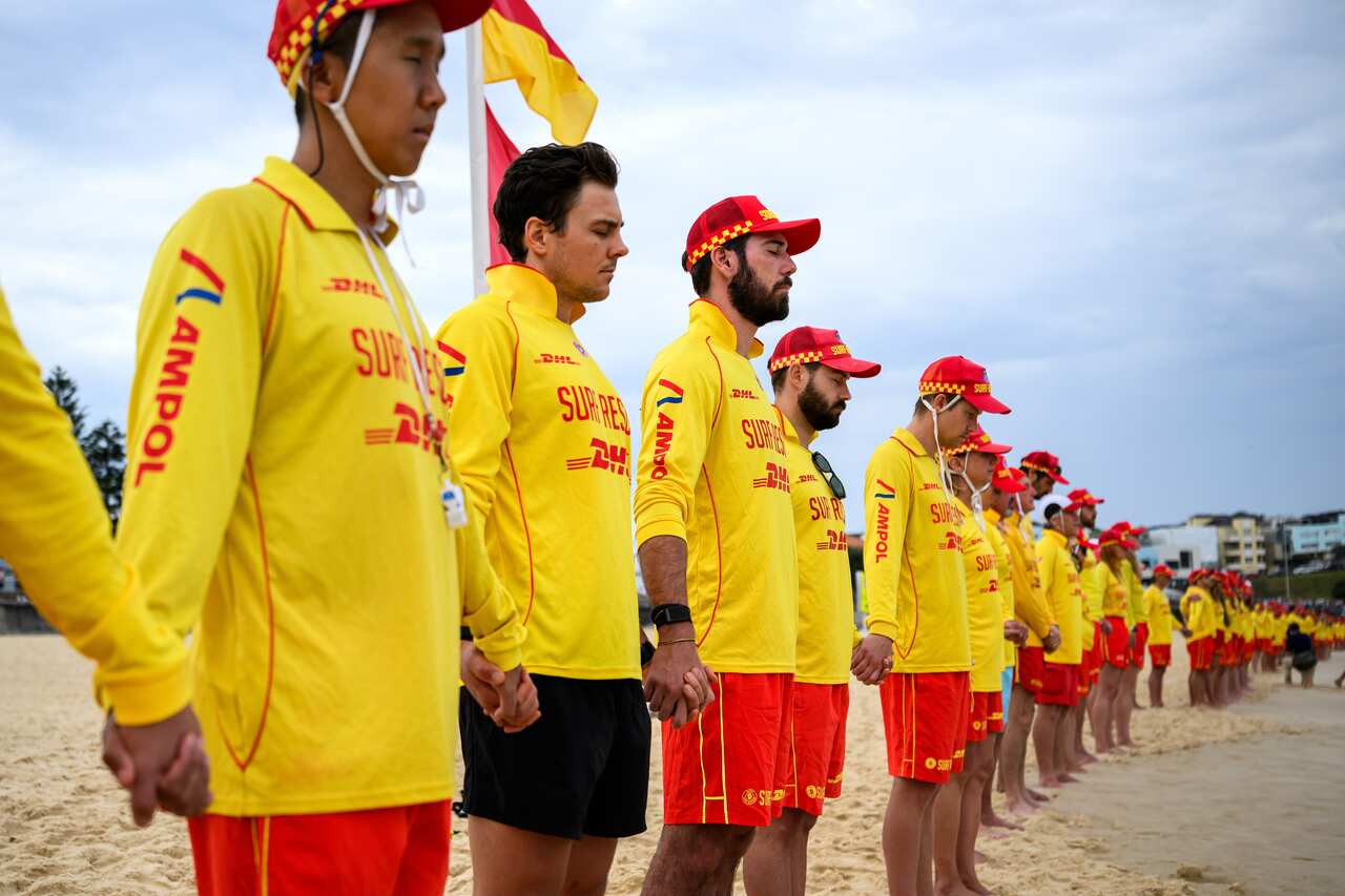 Lifesavers in yellow stand on a beach in a row, holding hands.