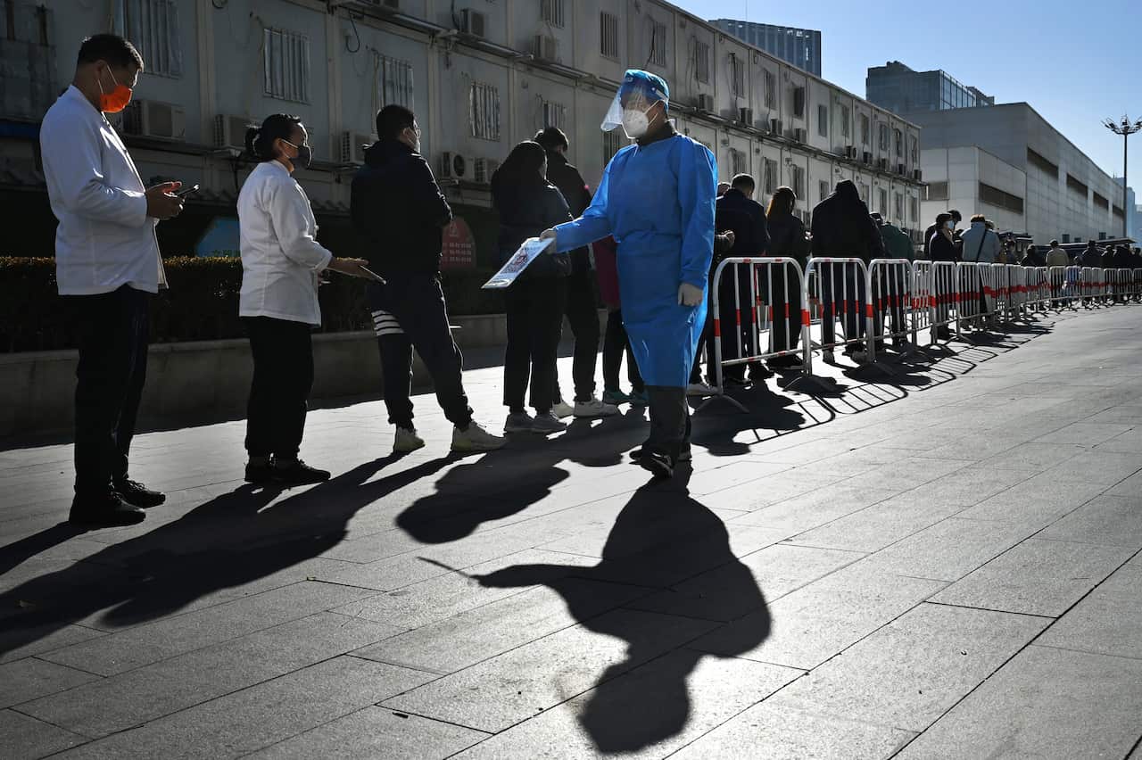 Person in blue medical gown walks past a line of people waiting to be tested.
