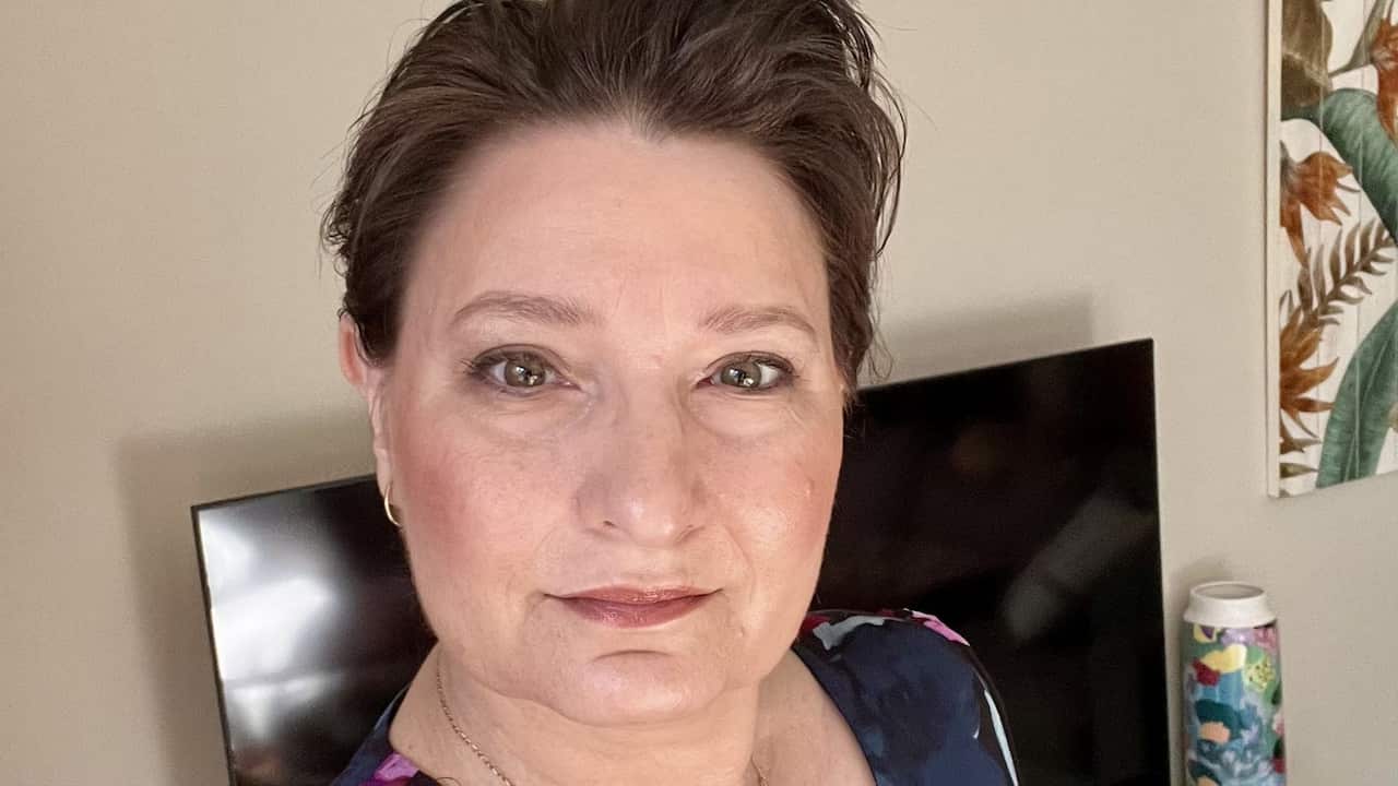 a close up selfie of a middle aged woman with makeup on taken in a living room with a tv in the background