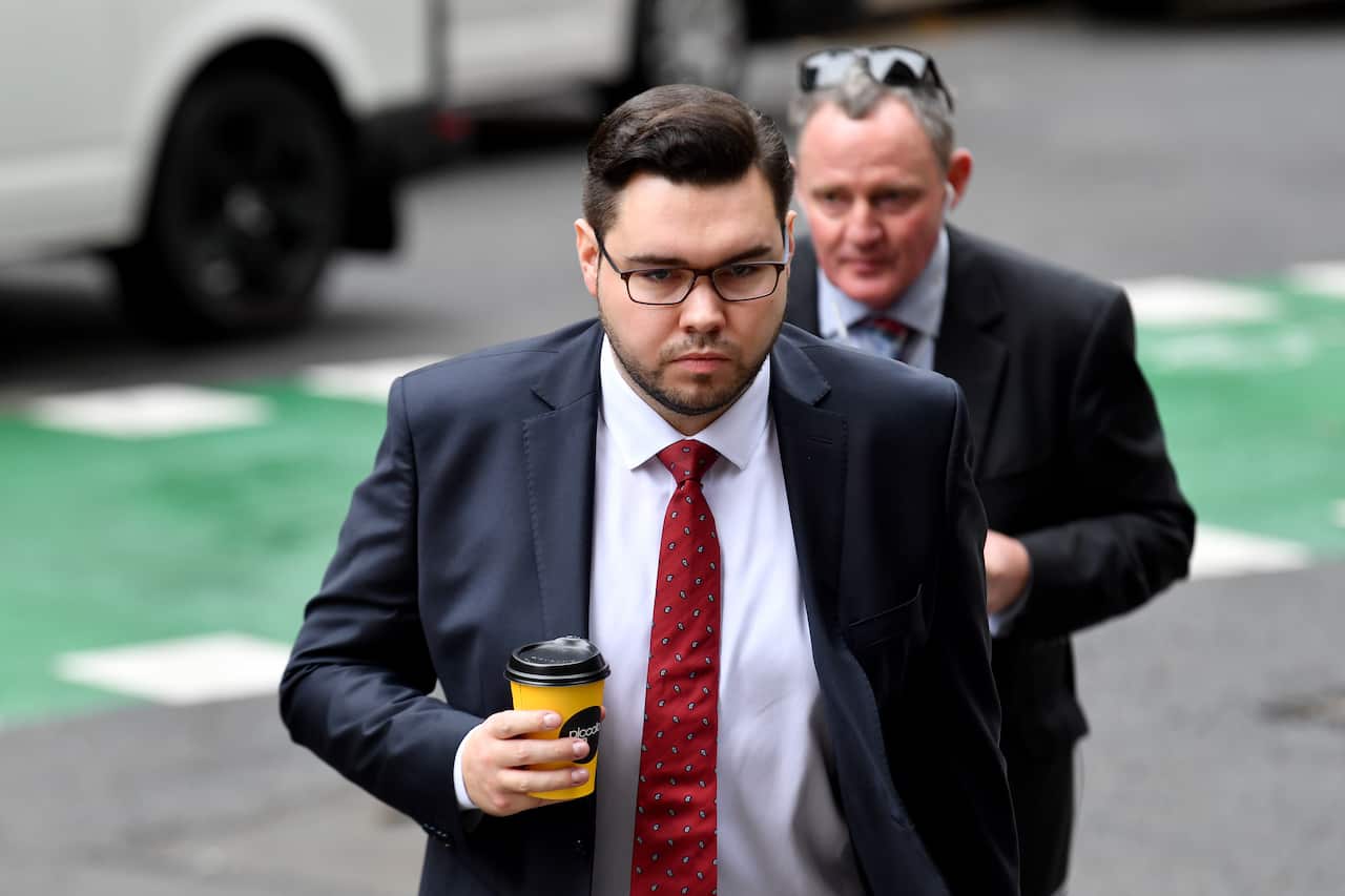 A man wearing a suit, tie and glasses walking outside and holding a takeaway coffee cup.