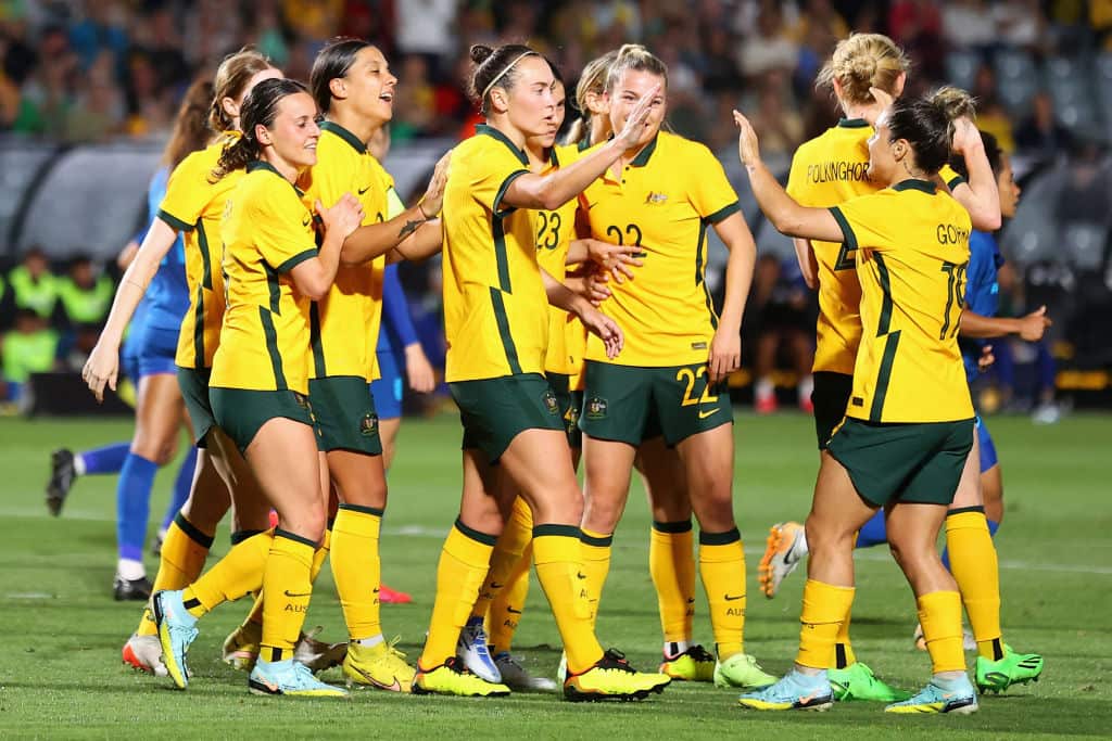 Women footballers wearing yellow jerseys, green shorts and yellow socks celebrate a goal