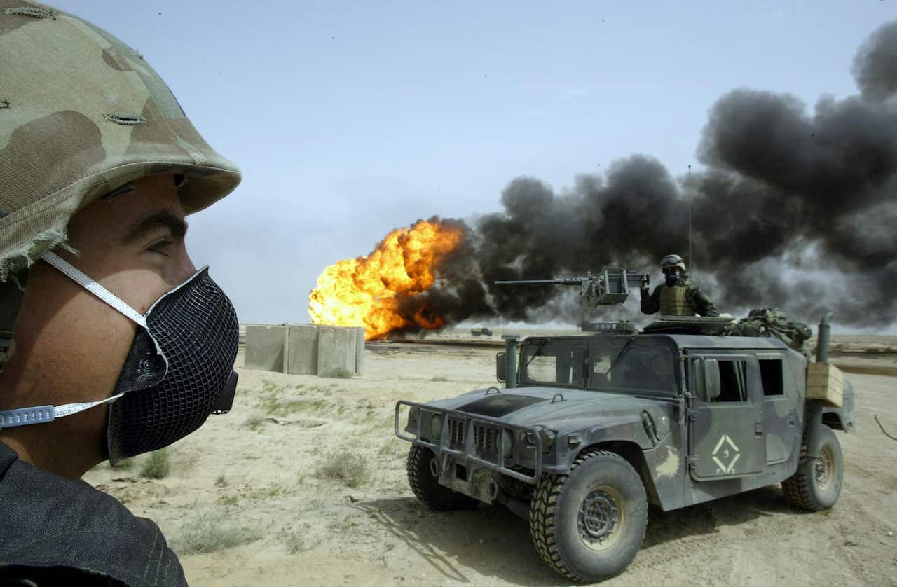 A United States soldier standing outside while another is in an armoured vehicle. A burning oil well is in the background.