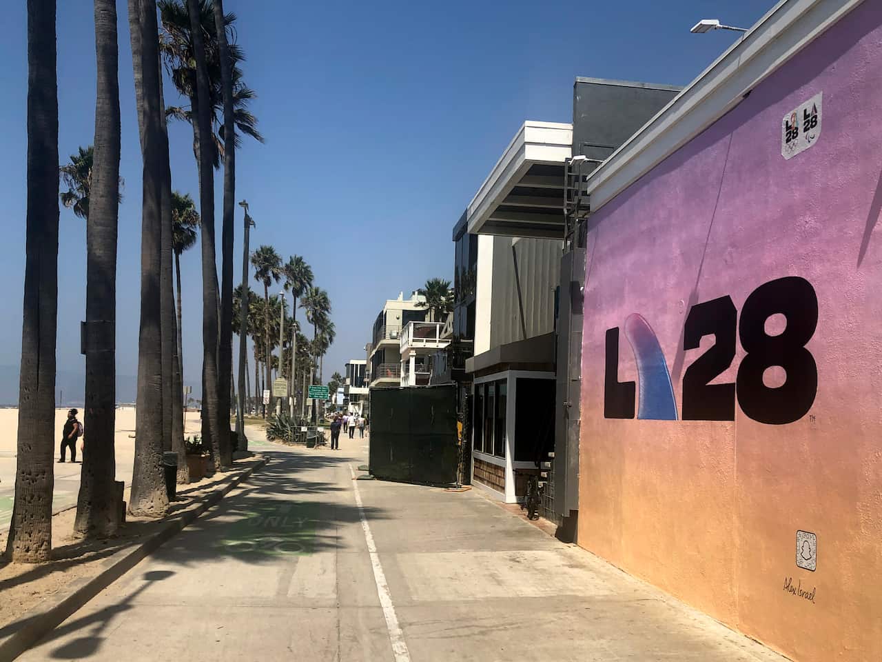 A wall with a LA28 logo on it next to a row of palm trees and a beach