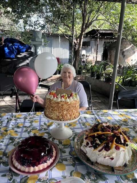 Elderly woman sitting behind 3 cakes on a table.