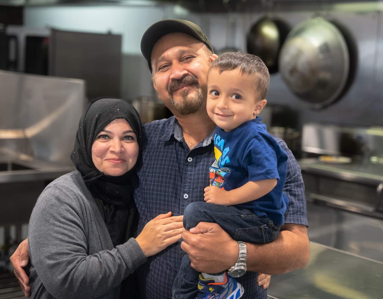 A woman in a grey shirt and a man in a blue shirt stand holding a small boy in a kitchen.