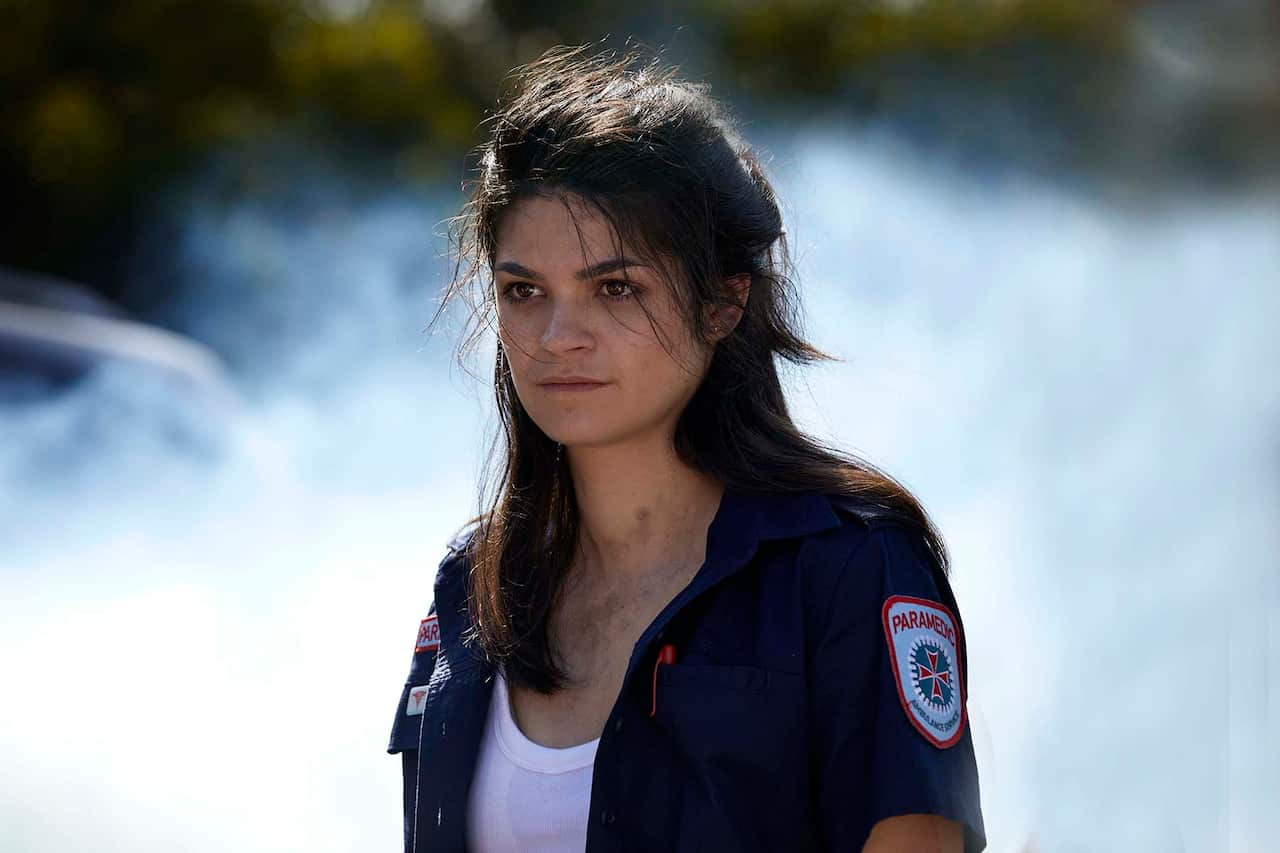 A head and shoulders shot shows a young woman in a paramedic uniform, standing in front of billowing smoke.