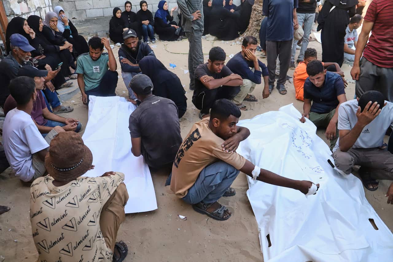 Men kneel beside two large white body bags. 