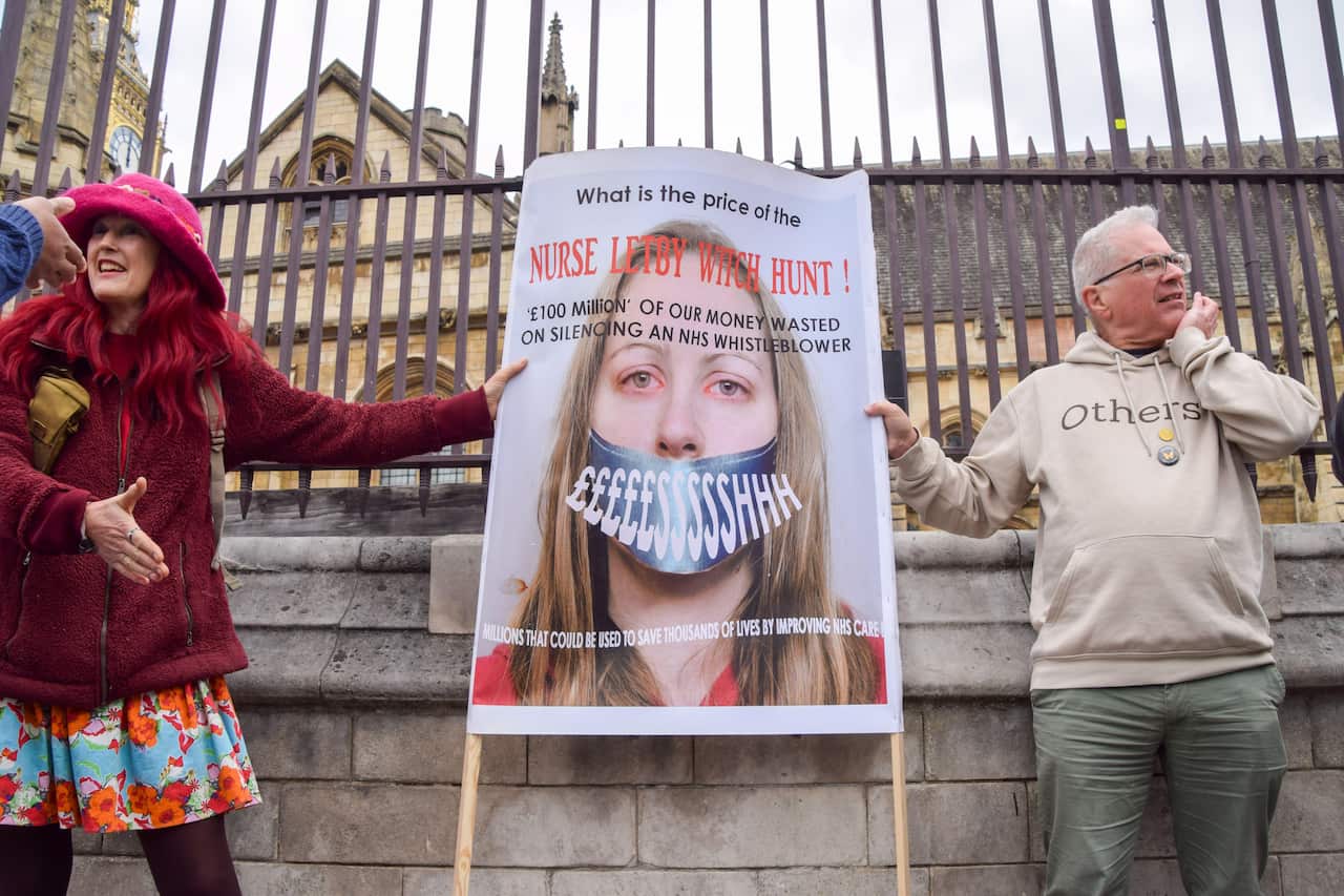 Two demonstrators outside a wrought-iron gate hold up a poster featuring a woman's face taped shut with text protesting a "Nurse Letby Witch Hunt."