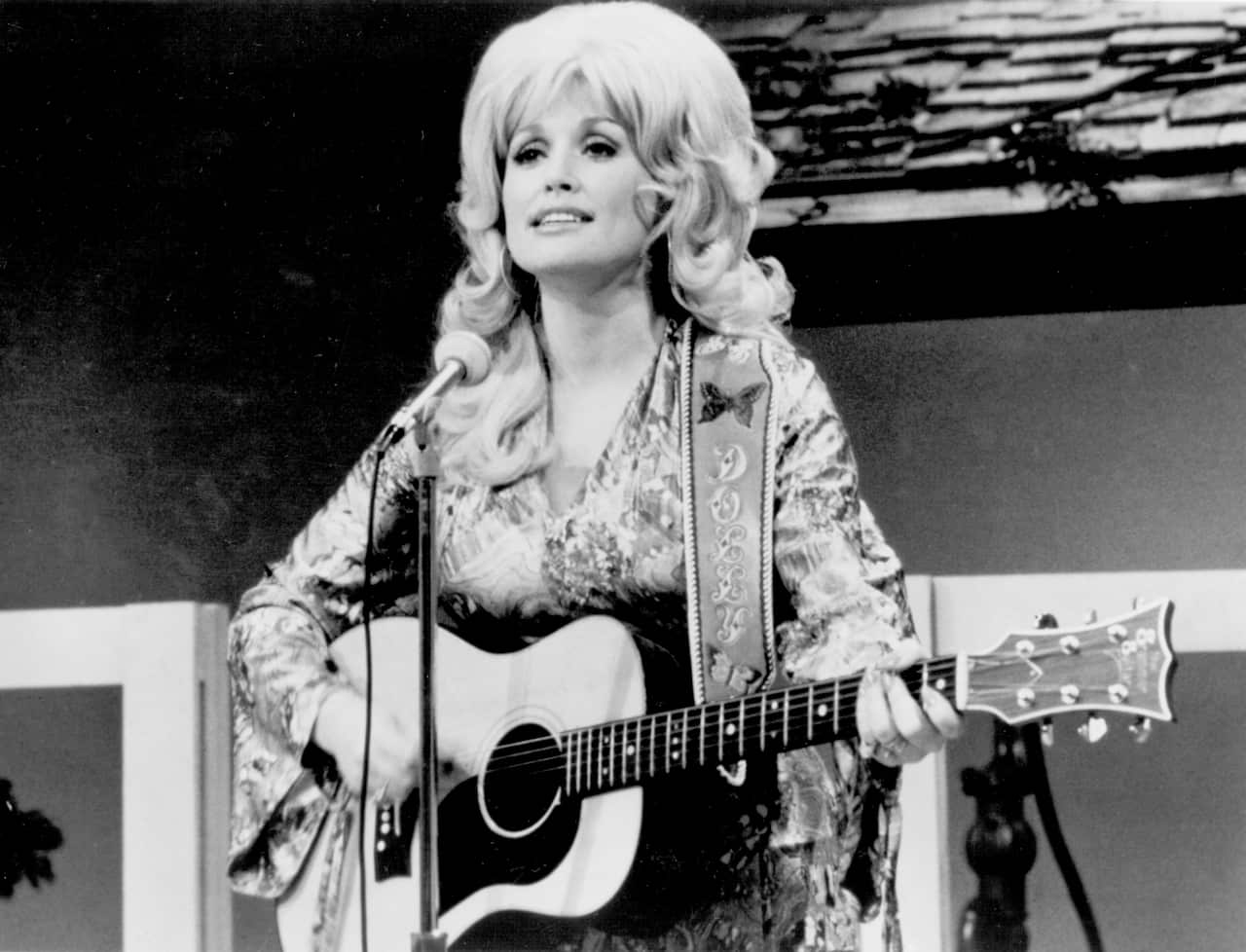 Black-and-white photo of a performer with a large hairstyle playing an acoustic guitar and singing into a microphone on stage.