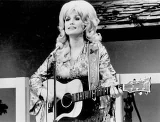 Black-and-white photo of a performer with a large hairstyle playing an acoustic guitar and singing into a microphone on stage.