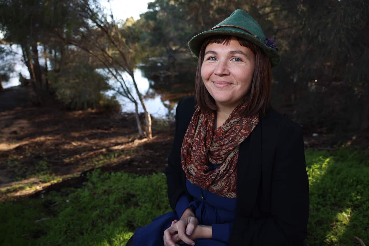 A woman in her late 30s looks to the camera, with a river bank and shaded trees behind her.