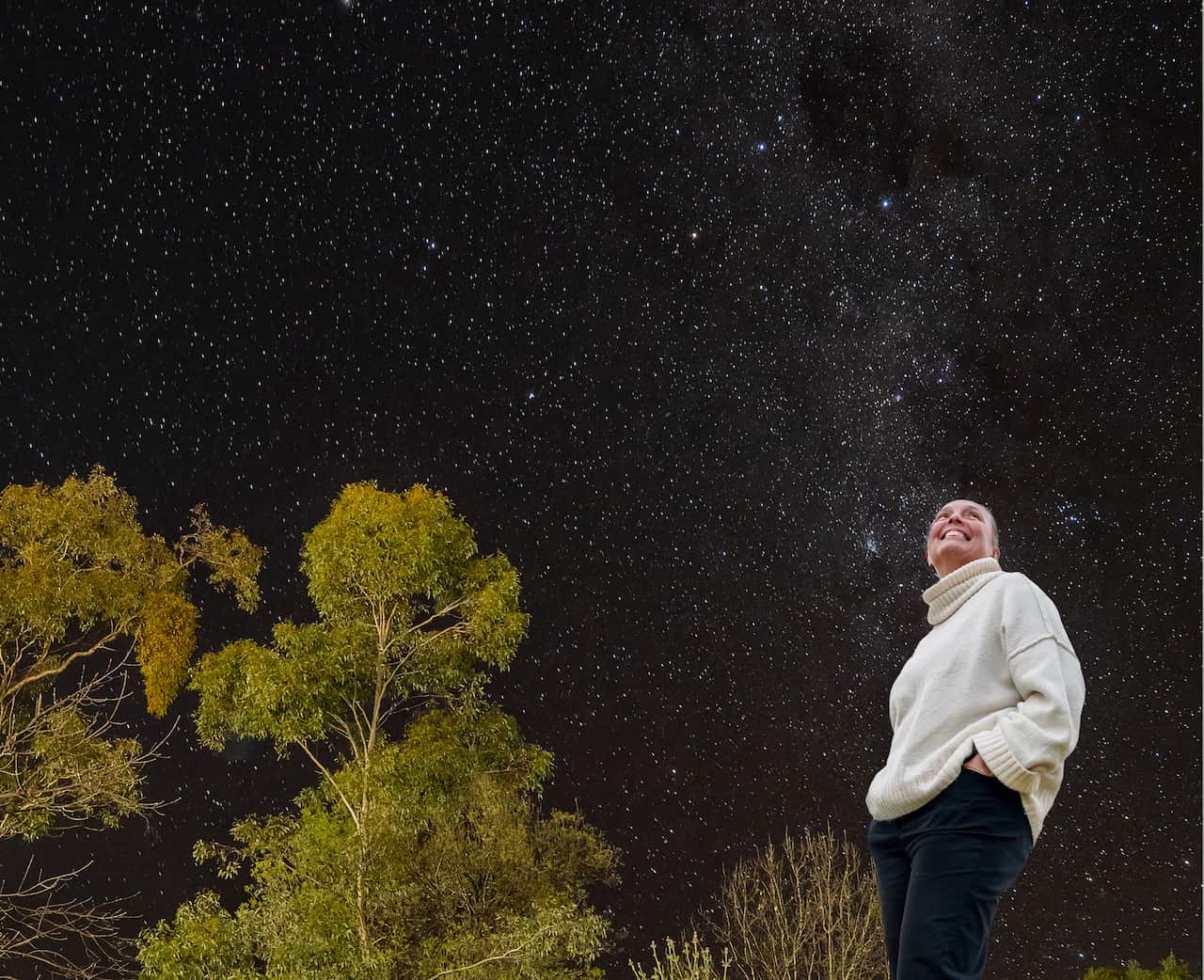 A woman smiles as she looks up at the stars. The photograph is taken from a low angle, with trees and the Milky Way in view.