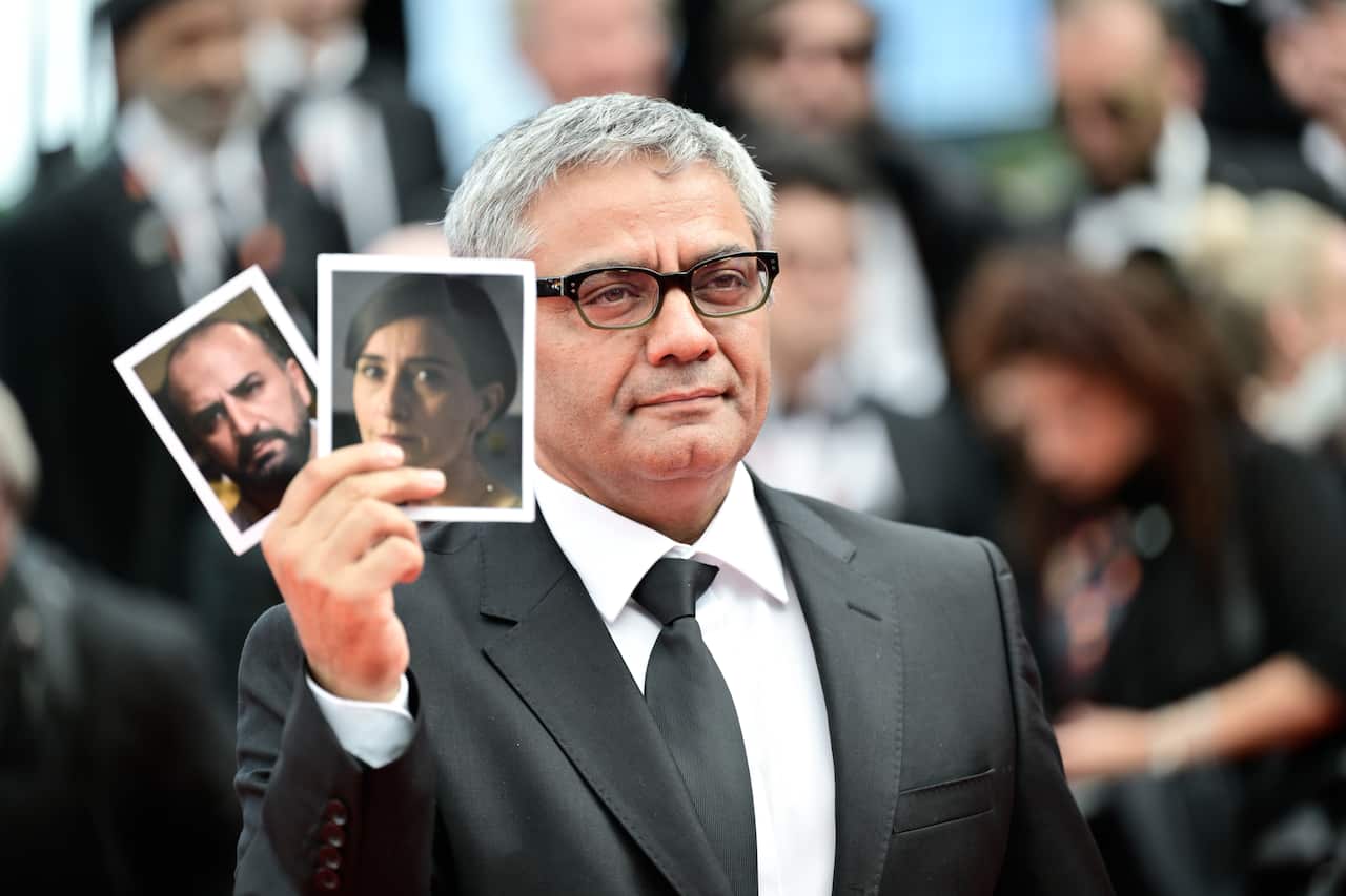 A man in a black suit and tie holds up two pictures - headshots of a man and a woman
