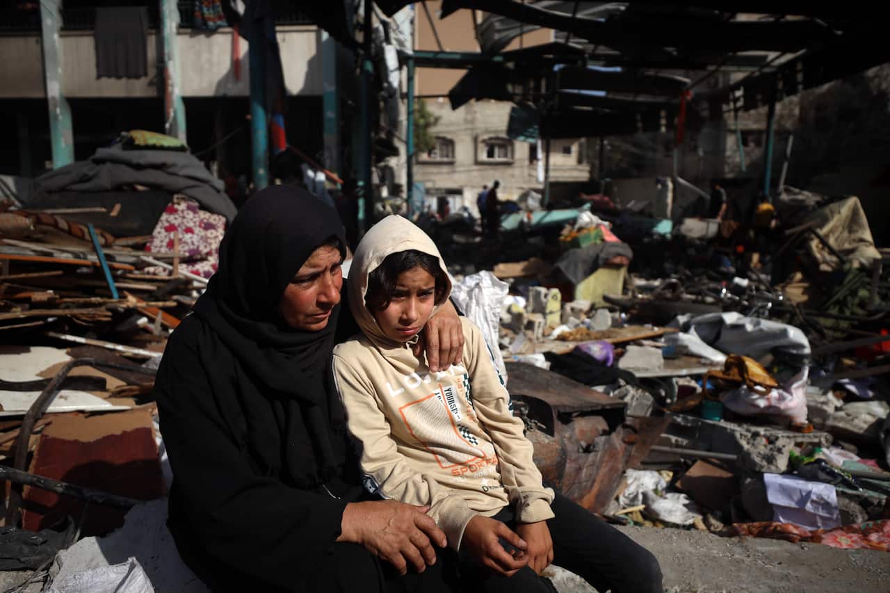 A child sitting on a woman's lap. Behind them are destroyed buildings and rubble.