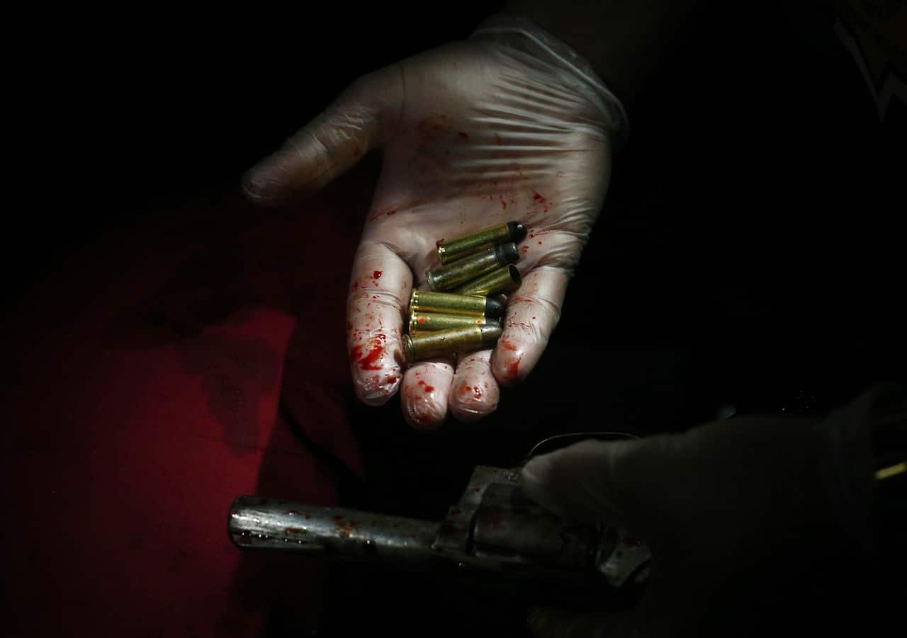 A hand wearing a glove with blood on it, holding bullets and a pistol.