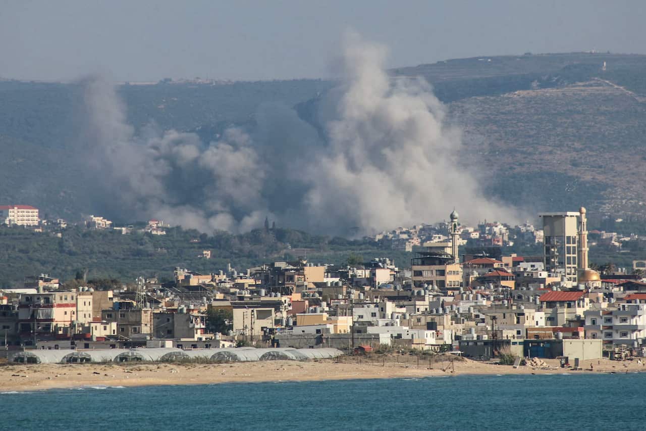 A shoreline with buildings and mountains in the background. Smoke can be seen rising.