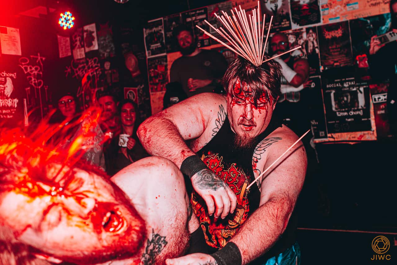Two men with skewers piercing their heads, covered in blood, are wrestling each other as a small crowd watches.