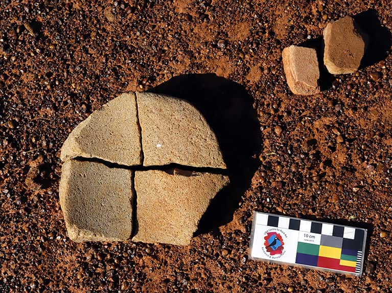 A broken grinding stone sitting on the red earth with a ruler placed nearby.