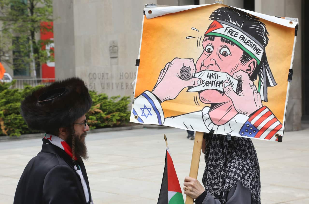 A person holding a placard with an image of a person with a 'Free Palestine' headband and a piece of tape over their mouth which reads 'antisemitism."