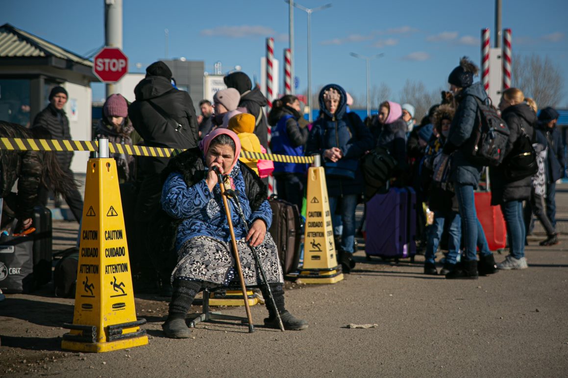 An elderly woman sits on a chair in front of a line or refugees from Ukraine at the Moldovan border.