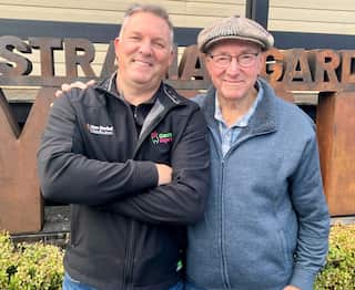 A man wearing a navy sweater stands next to an older man wearing a peaked cap.