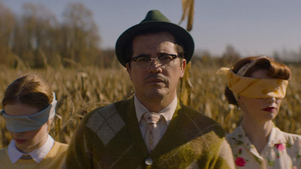 A man stands in a corn field with a woman and young girl blind folded on either side of him.